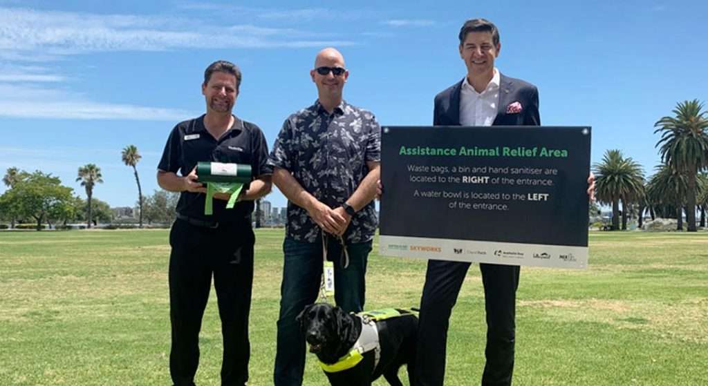 Image shows a group photo with David Vosnacos, Guide Dog User Eric Seery with Guide Dog Sundae lying on the grass and Perth Mayor Basil Zempilas holding a sign.