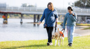 Mum, child and Autism Assistance Dog walking on grass along river.