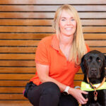 Guide Dog Mobility Instructor kneeling next to black Guide Dog in harness.
